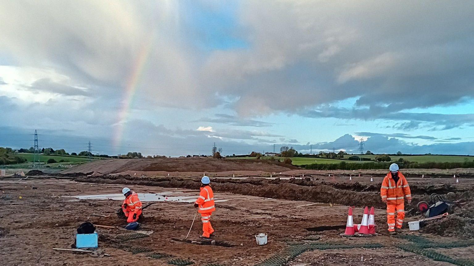 A rainbow leading towards a large scale archeological dig with people in orange high viz suits and hard hats working