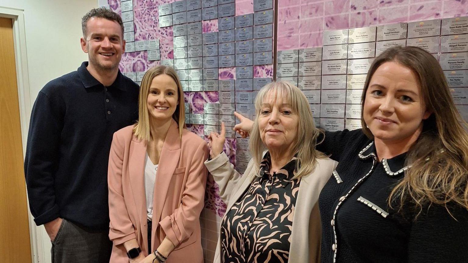 One man and three women stood against a wall, all looking at the camera. Two of the women are pointing to a silver plaque on the wall. 