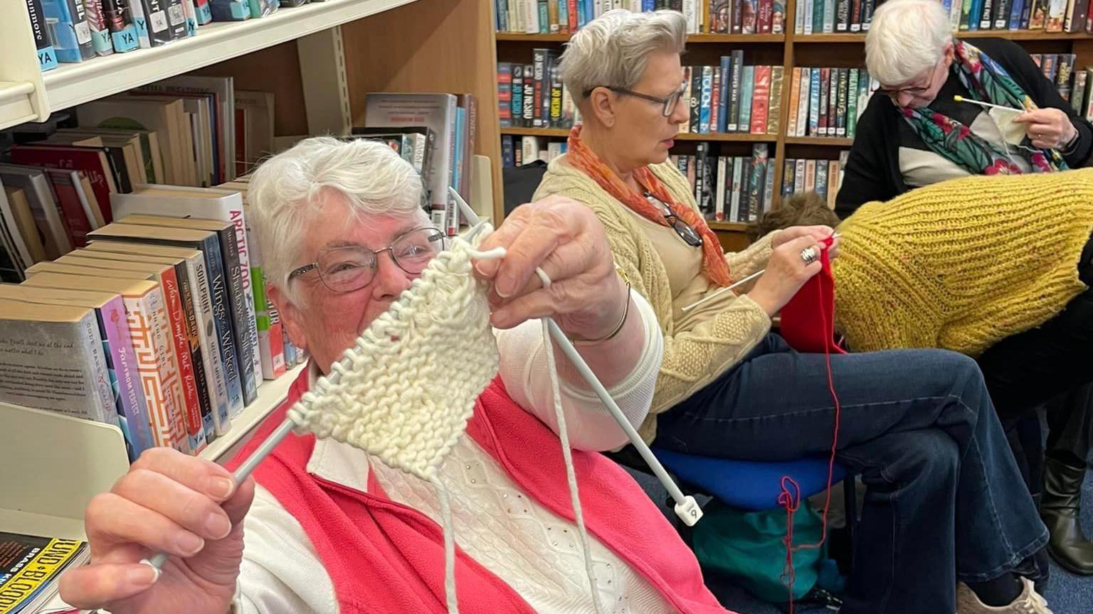 A group of three ladies are sitting on chairs in the library knitting, one of them has pointed her knitting needles and a small piece of knitted fabric and the camera