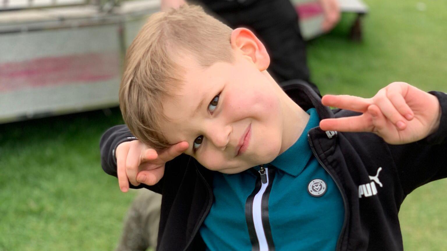 A little boy, who has his head to one side and is smiling. He is wearing a teal top and black hoodie. He is standing in a field and holding both his arms up near his face so his elbows are bent and he is making the peace sign with both of his hands. He has short, light brown hair.