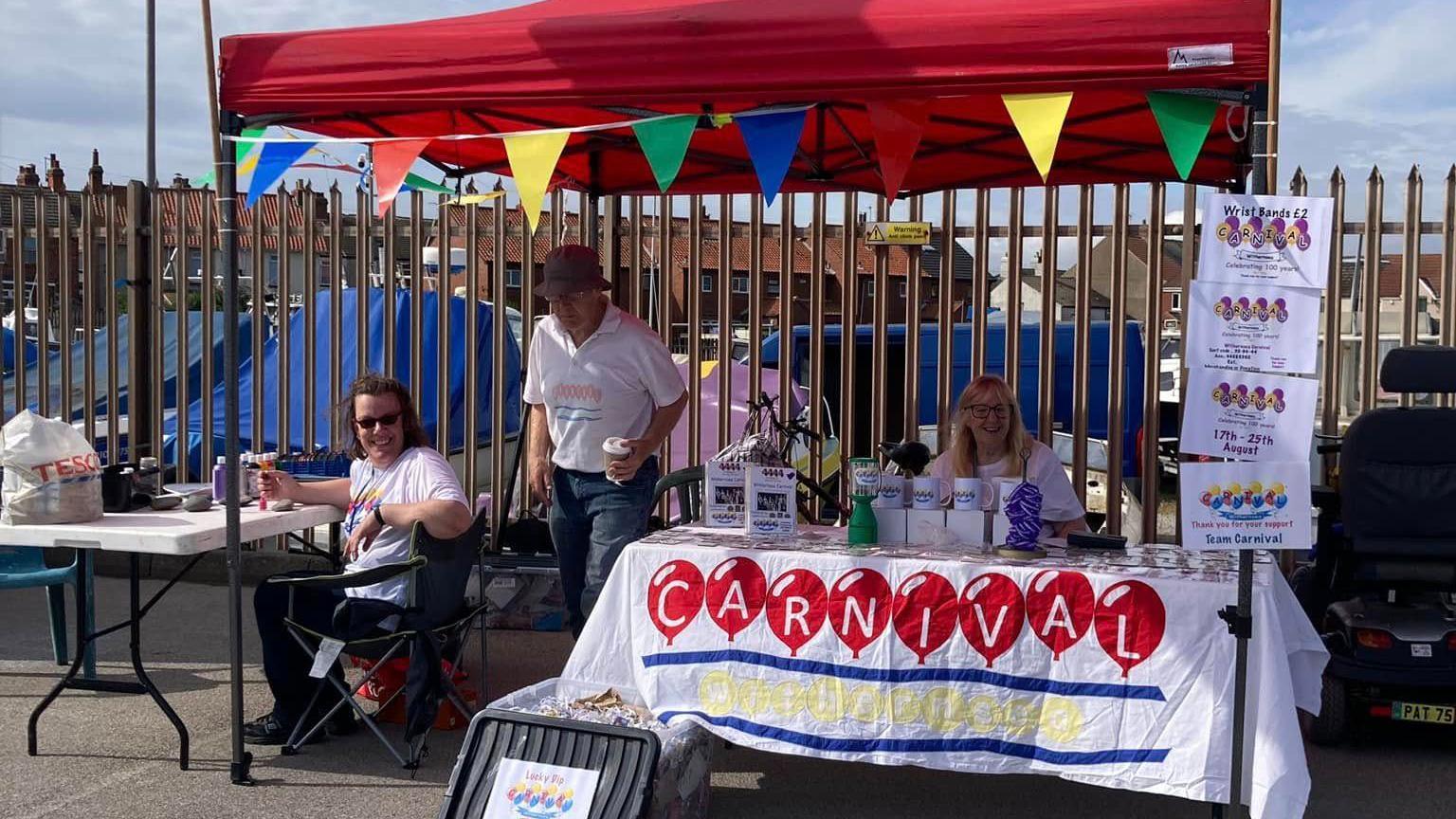Three people sitting under a red canopy stall decorated with colourful bunting and flags. A table displays a sign reading “carnival” in capital letters.
