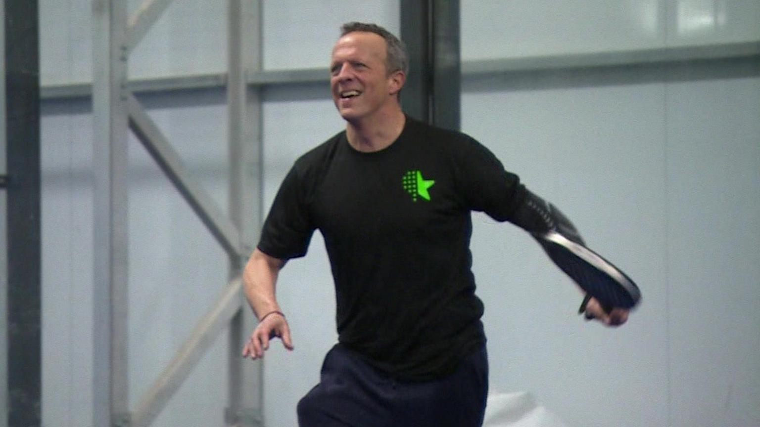 Andy Knee playing padel, wearing a black t-shirt and tracksuit bottoms, holding a racquet.