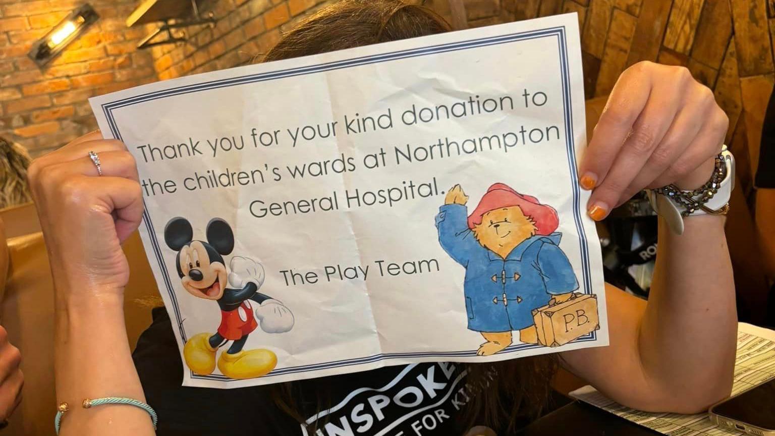 A volunteer holds up a sign which reads: "Thank you for your kind donations to the children's wards at Northampton General Hospital - The Play Team." The poster is decorated with an image of Mickey Mouse and Paddington Bear.