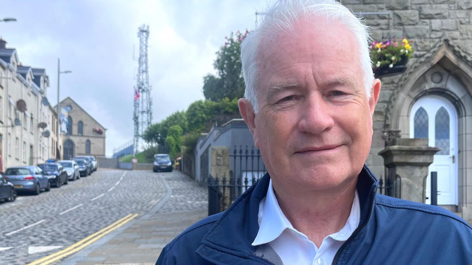 Man with white hair, wearing a white shirt and navy coat.
In the background is a street lined with cars and the metal mast. 