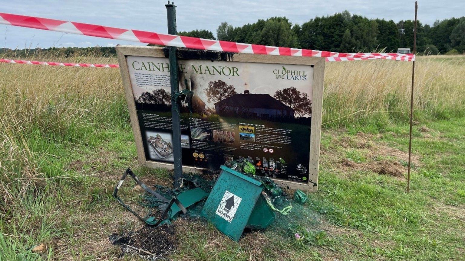 Vandalised and charred information board in a grassy field, surrounded by red and white tape. The ruins of a destroyed bin are in front of it.