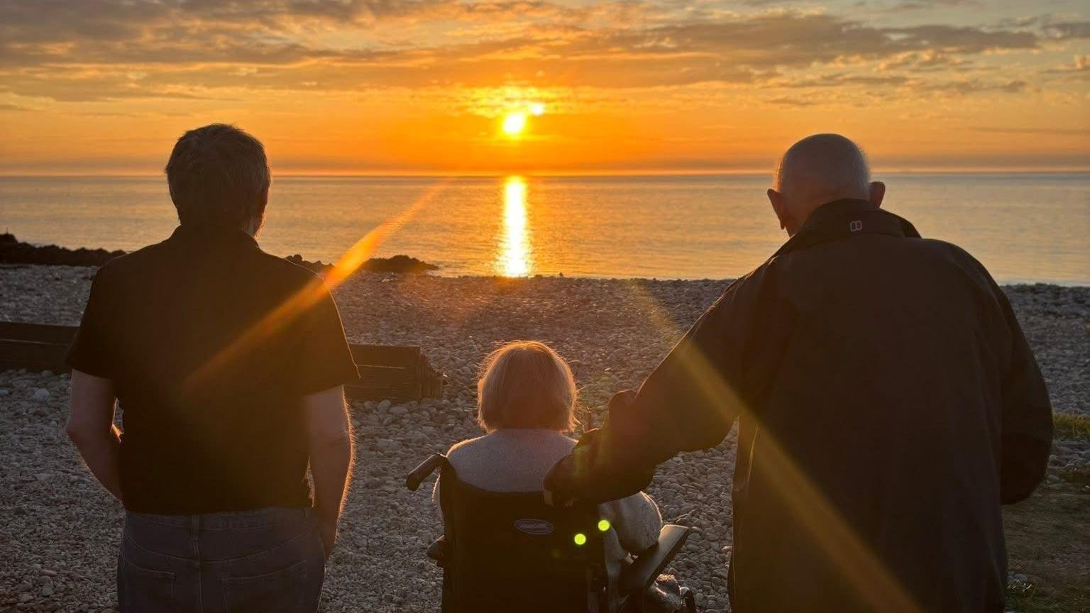 the backs of two men and a woman, who is in a wheelchair, over looking the water with an orange sun setting in the background.