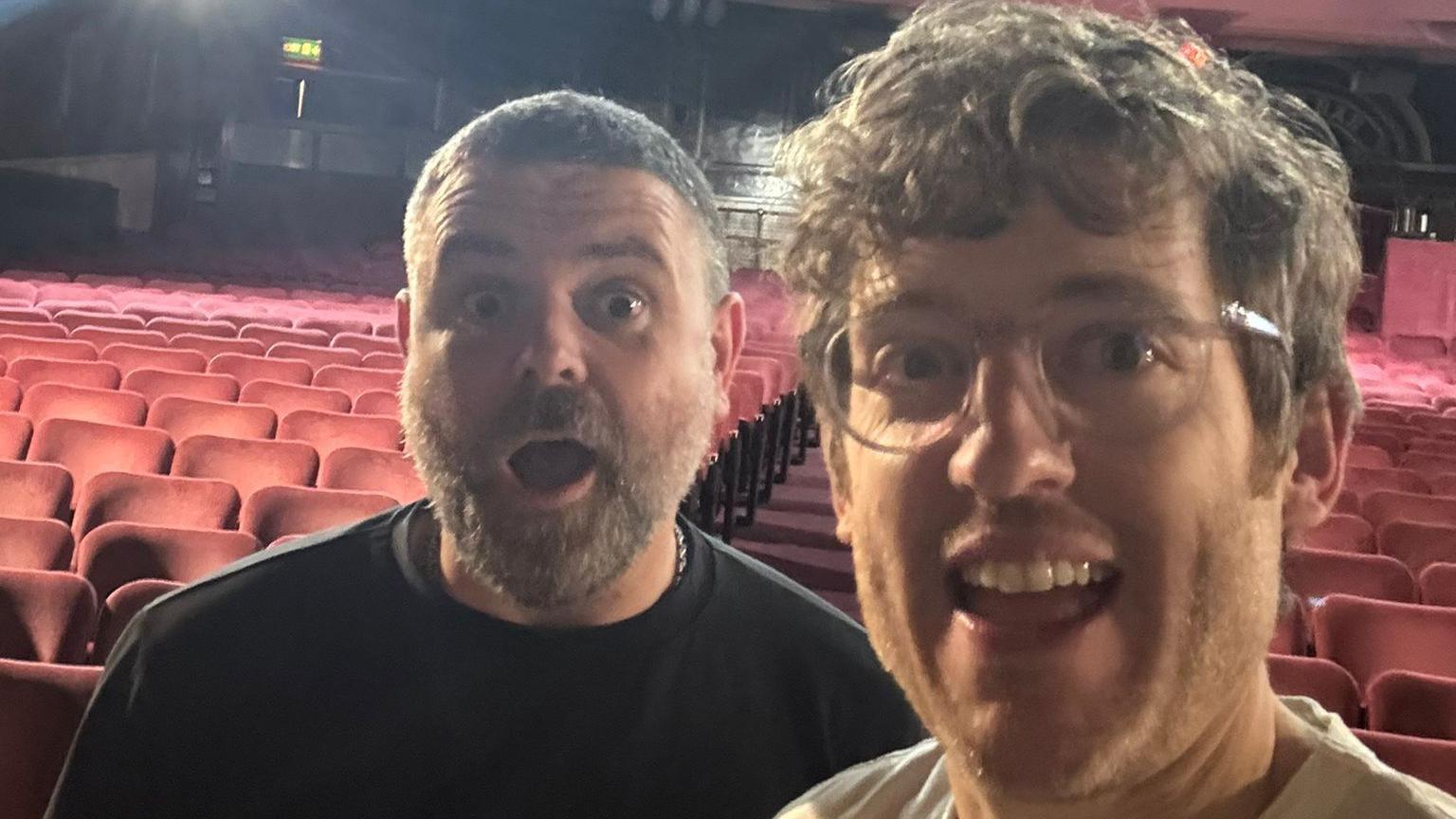 Geraint and Elis in an empty London Palladium both smiling with a fun open-mouthed expression at the camera. Geraint wears a black jumper with a silver chain and Elis wears a cream t-shirt and translucent-rimmed glasses.