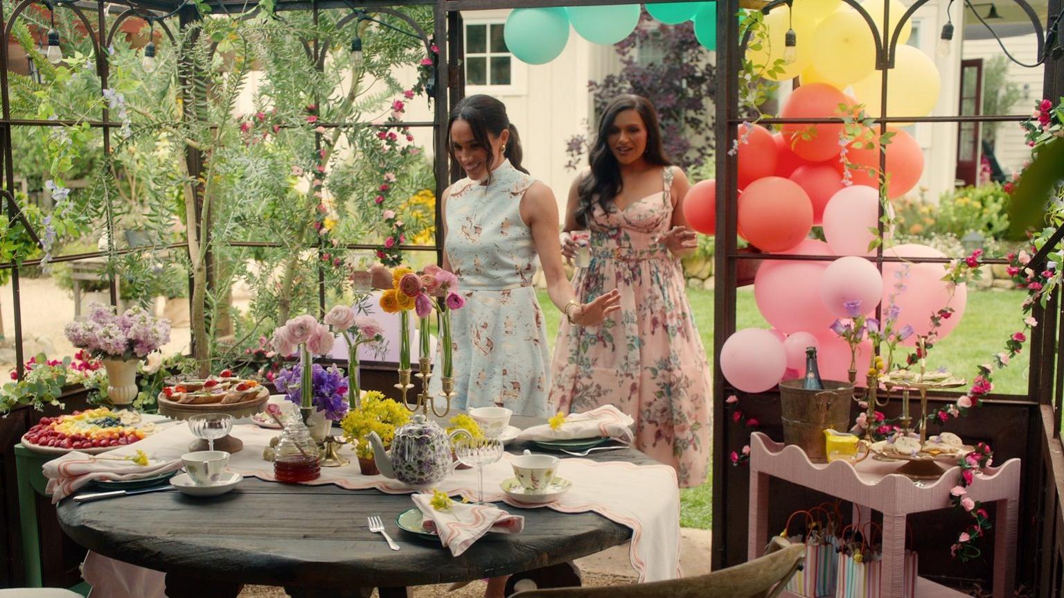 Meghan, seen with actress Mindy Kaling, both wearing floral dresses and walking into a garden room made up for a tea party