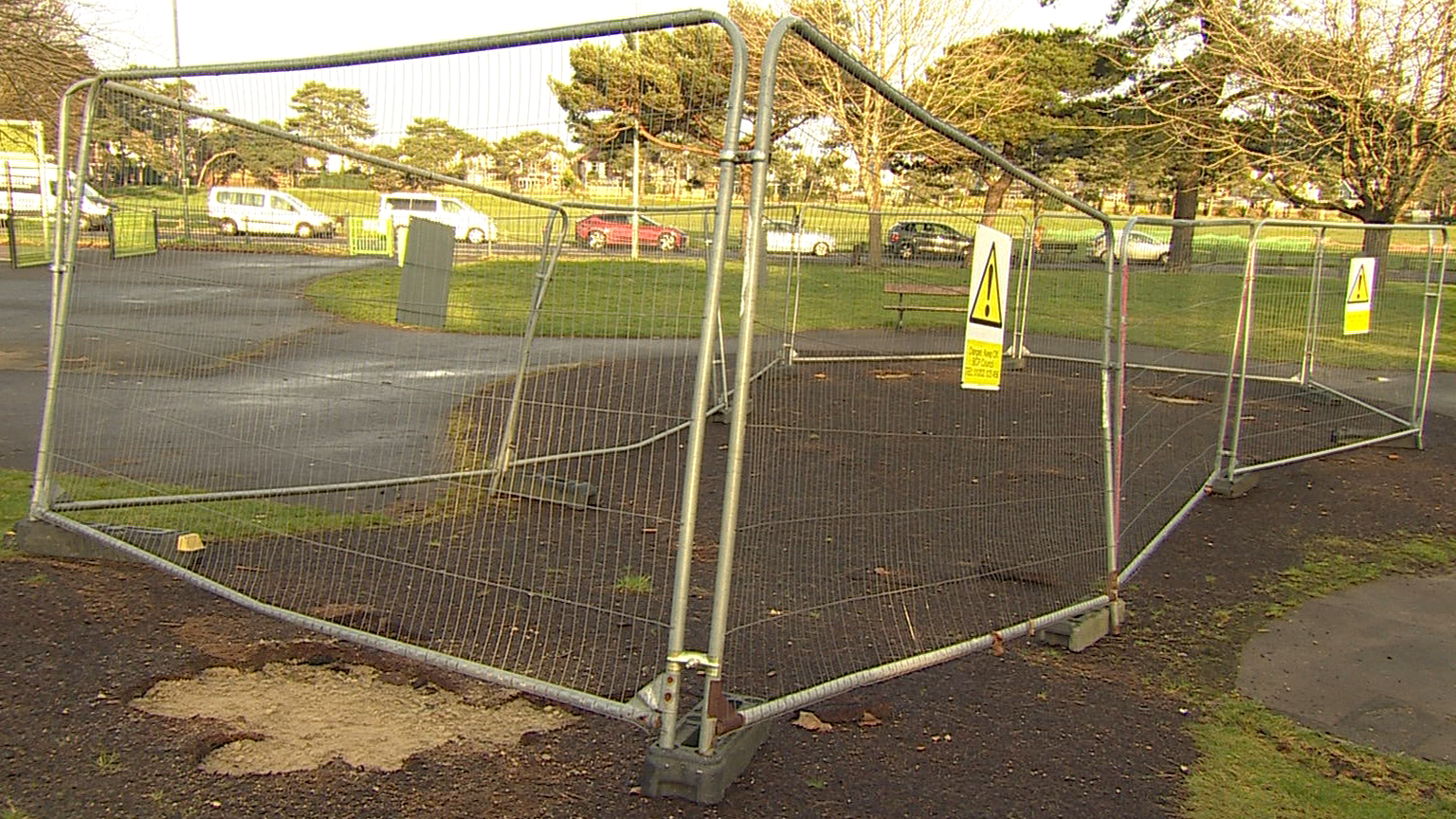 Fencing sealing off an area where wooden play equipment stood until recently. There are potholes and the area behind the fencing is closed to park users.