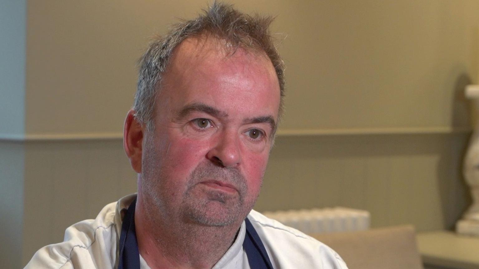 Richard Sim is sitting and speaking to an interviewer off camera. He has short dark grey hair and is wearing a blue apron. He looks serious.