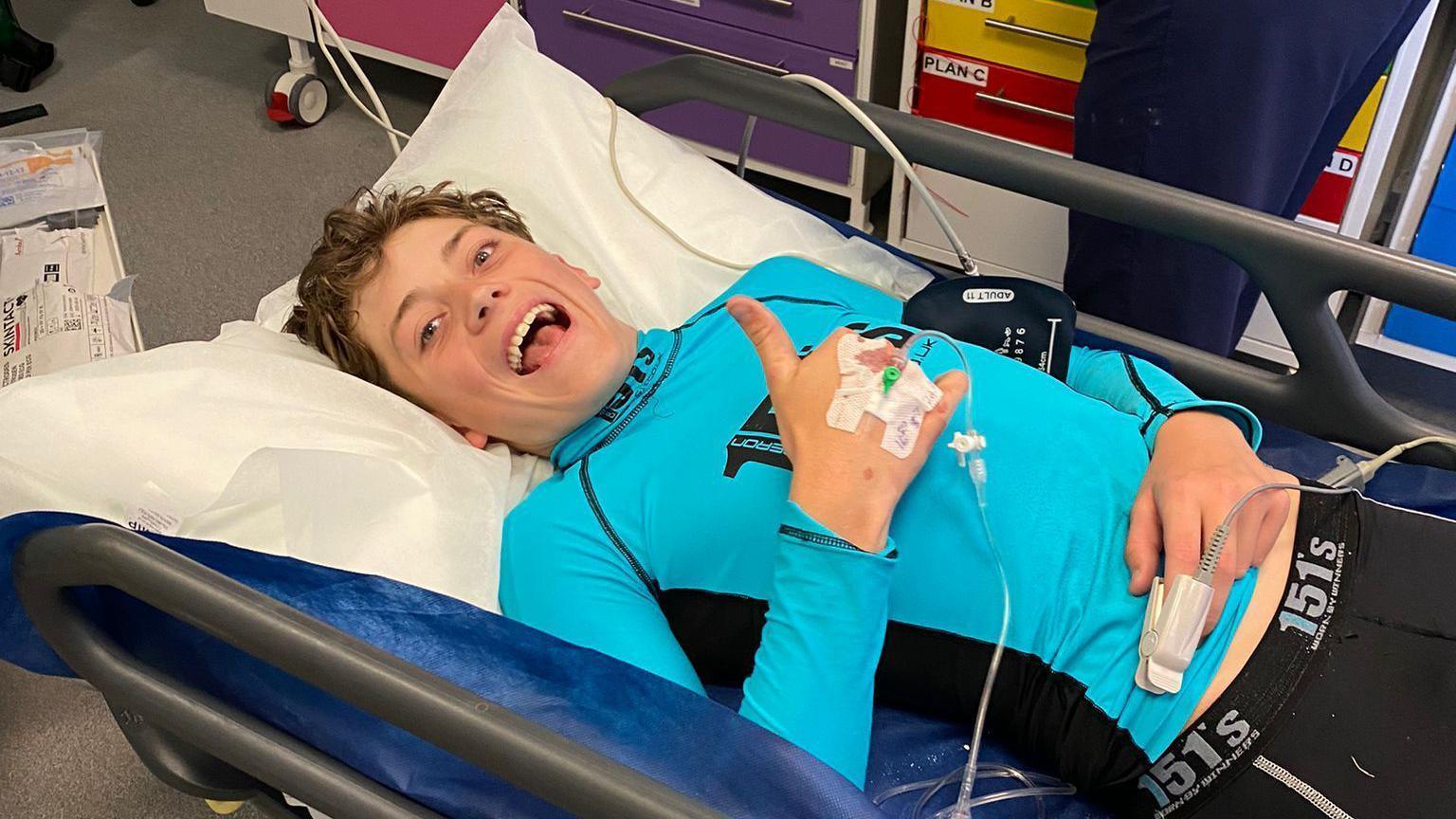 A smiling Charlie Huntingford on a hospital stretcher giving a thumbs up to the camera, wearing a blue long-sleeved vest top. 