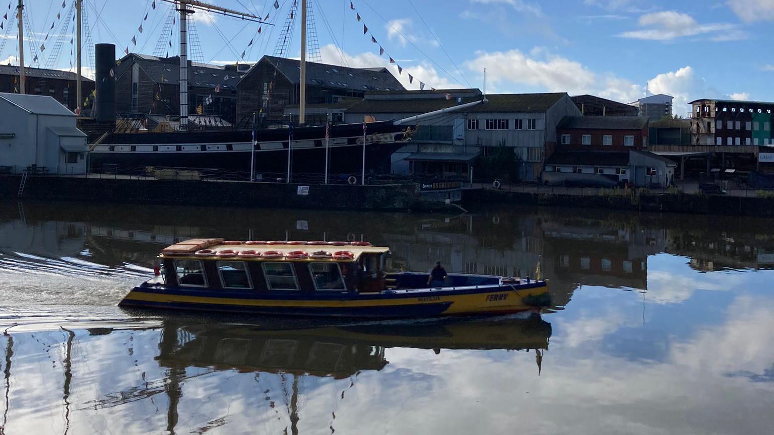 A blue and yellow ferry seen gliding past the SS Great Britain. The water is very still, with the reflection of the blue sky in it. The SS Great Britain can be seen in the background with warehouses behind it. 