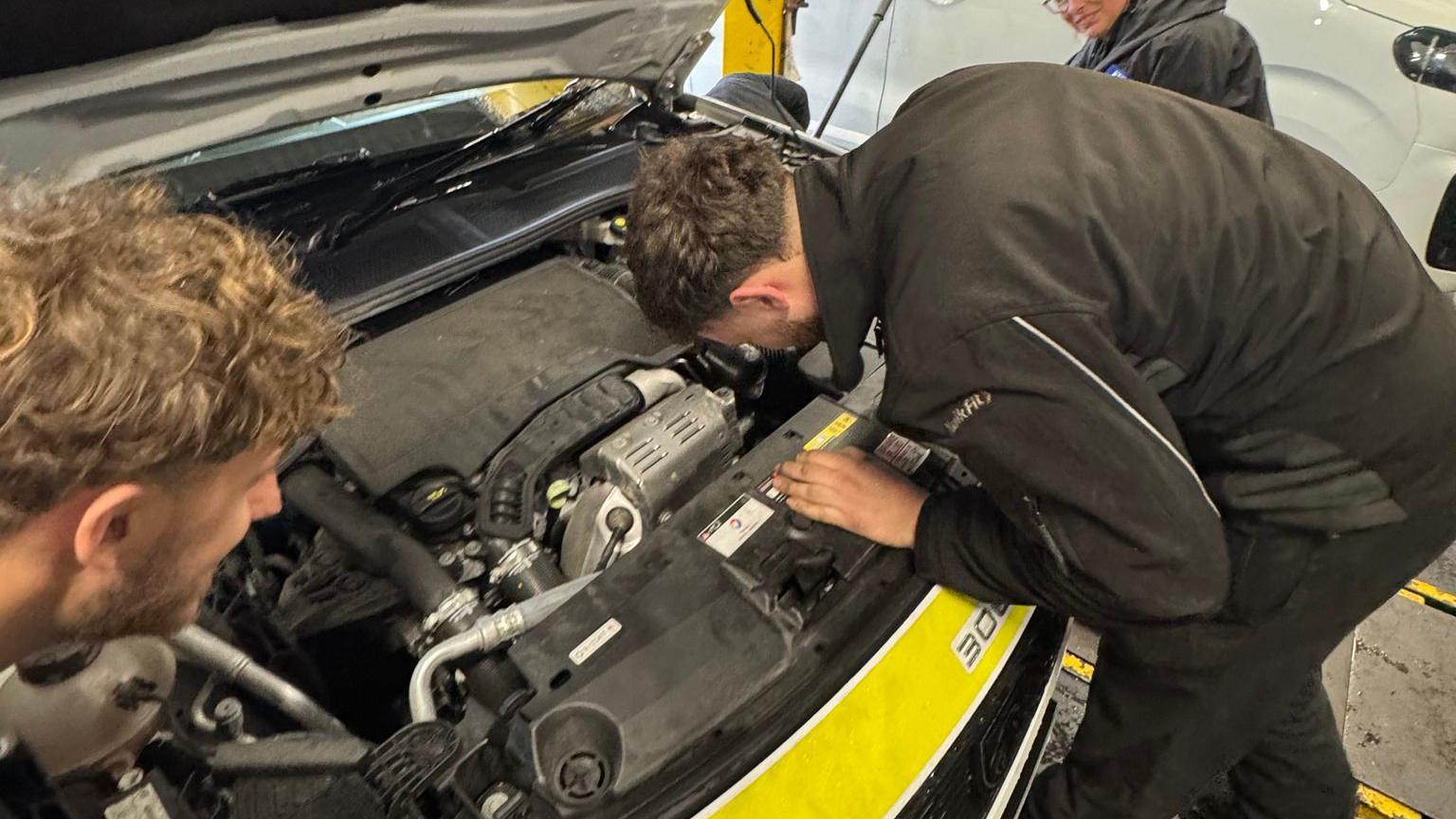 A picture of two mechanics looking into the engine of the police car. They are wearing black outfits and the front of the car is a bright yellow colour.