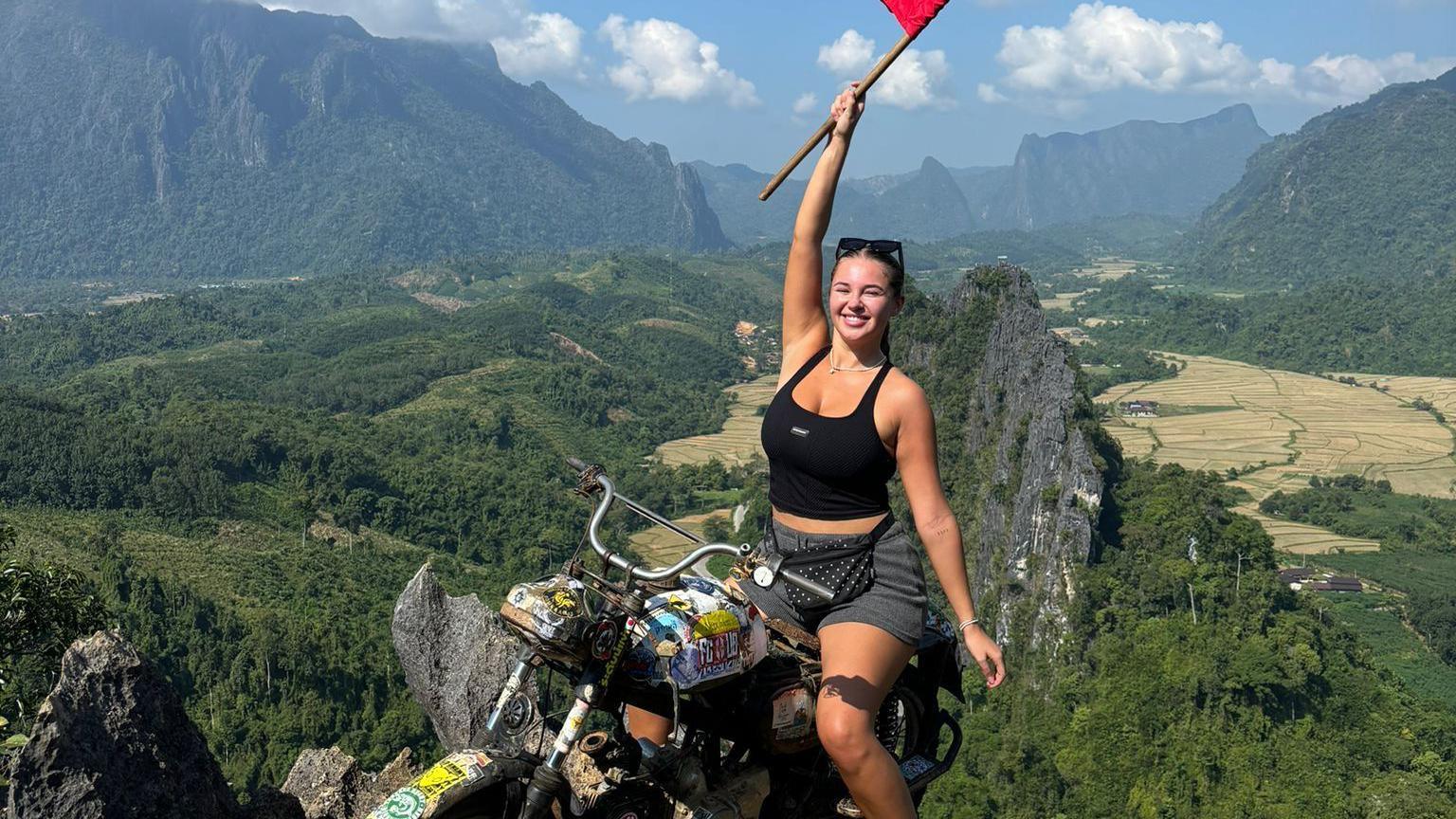 Natasha Moore, who wears sunglasses on her head and a black vest top with grey shorts and is pictured on a motorbike holding a Laos flag above a valley, in Vang Vieng