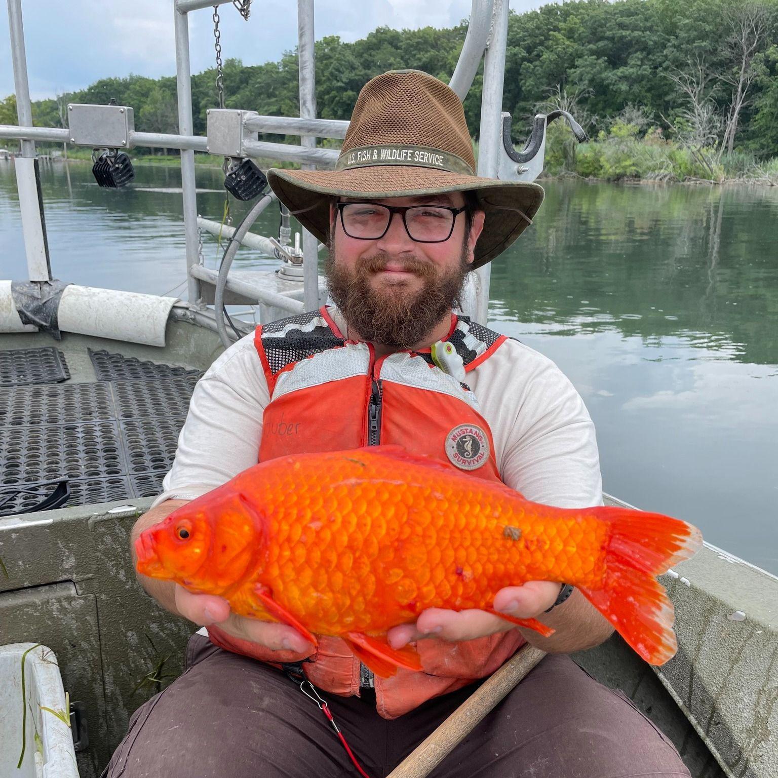 Man sitting on a boat holding a goldfish the size of an adult's male shoe up to the camera 