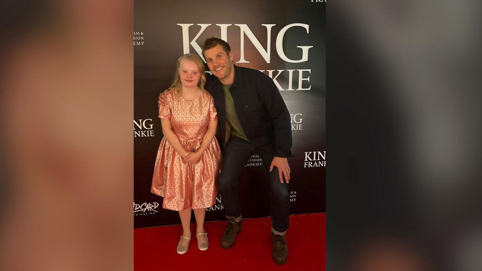 Claudia standing next to Peter Coonan. Claudia has long blonde hair and is wearing a pink dress with a pair of gold pumps. Peter is wearing a black jacket, green top, denim jeans and brown boots.