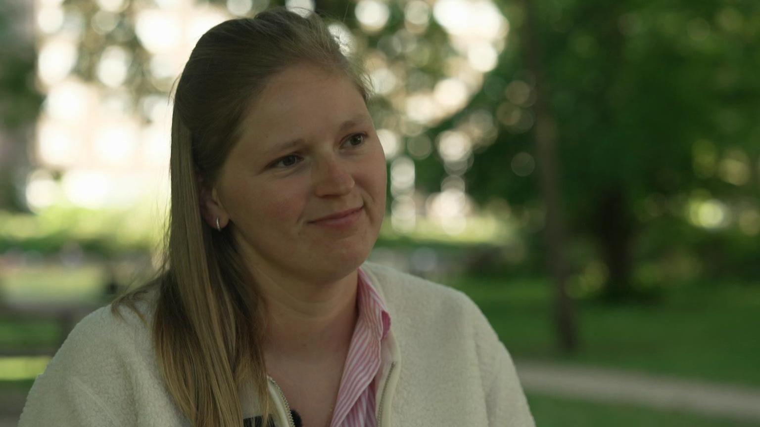 Charlotte, who has long light-brown hair and is wearing a white cardigan and pink and white striped shirt, is sitting in a park
