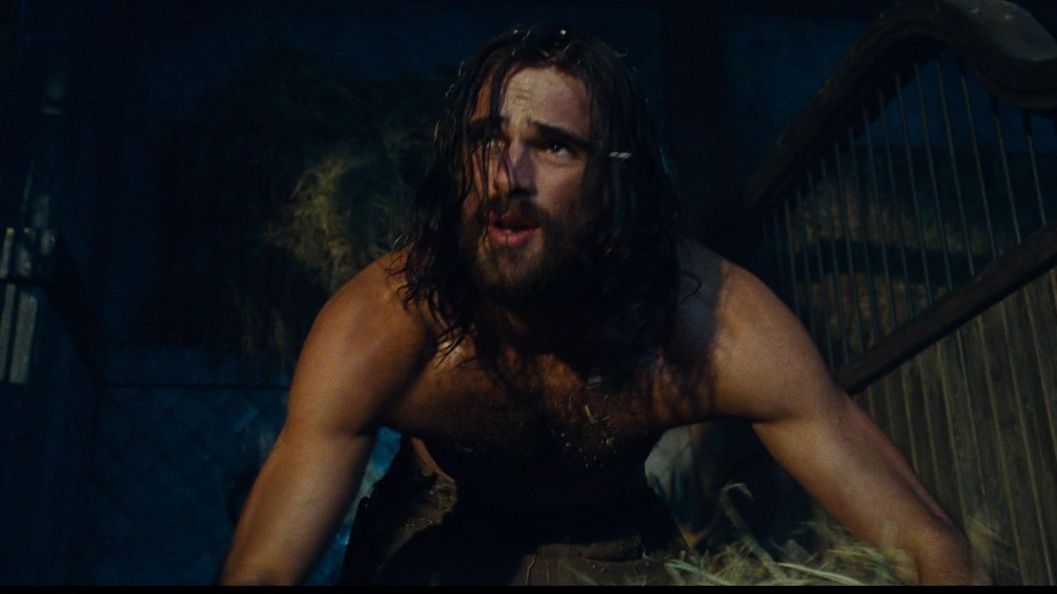 Jacob Elordi is pictured as Heathcliff in a still from the film, working in what seems to be a stable. He is topless, bending down to pick up straw, his chin-length dark hair falling across his bearded face.