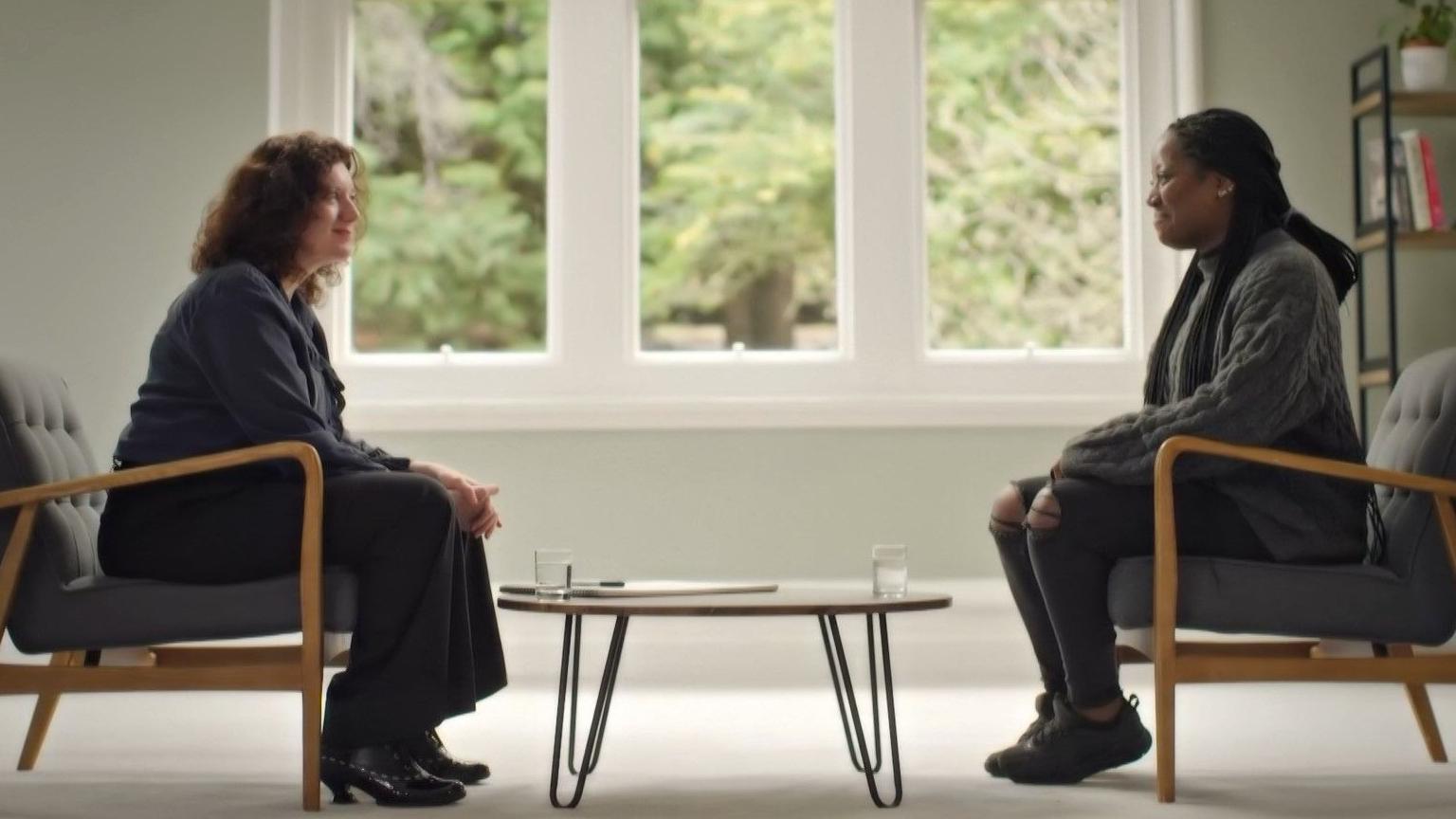 A white woman with brown wavy hair sat to the left of a low level coffee table. To the right is a black woman wearing a grey jumper looking a bit nervous. 
