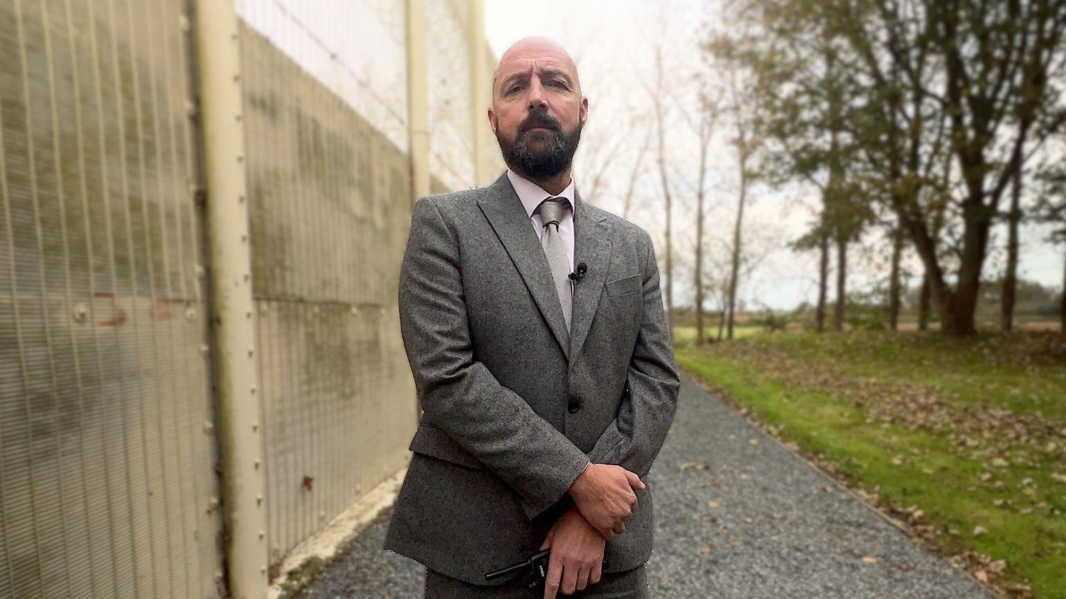 John De Carteret is wearing a grey suit with a white shirt and pale coloured tie. He has a thick dark beard and is standing in front of a concrete fence