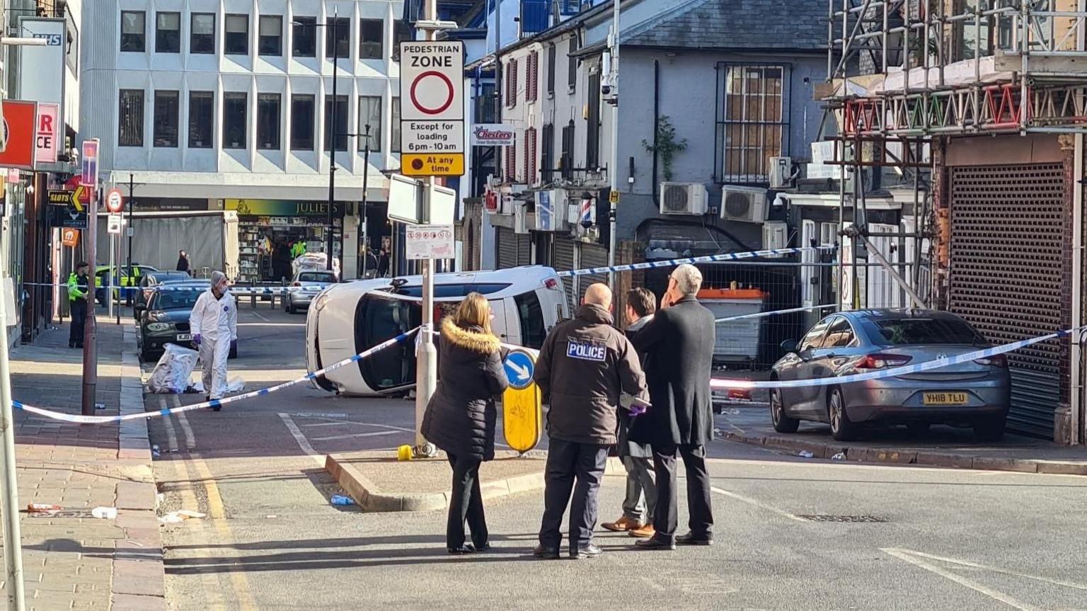 Four people, including a man with a black police jacket on, stand in front of blue and white police tape stretched across the road. Beyond the cordon is a crashed white car on its side. There is a police forensics officer in a white suit near the car.