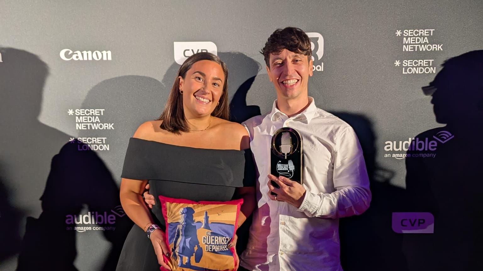 A woman and a man stand next to each other at the award ceremony. The woman, on the left, is wearing a black dress and is holding a squared-shape item which says The Guernsey Deportees. The man, on the right, is wearing a white shirt and is holding a trophy. The pair are smiling.