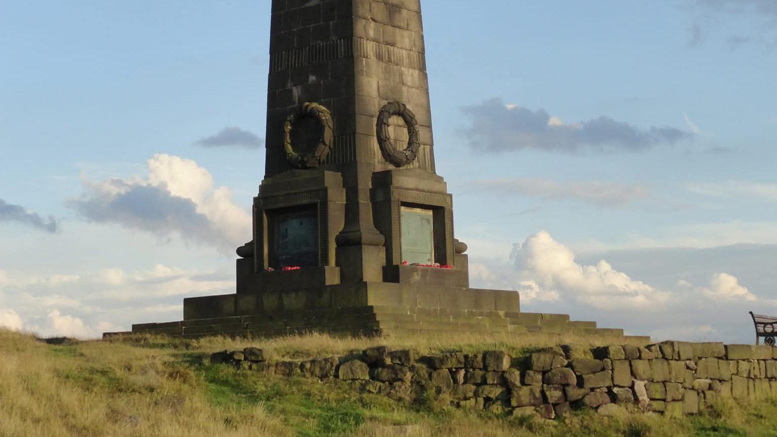 Dark stone base of the tapered obelisk on the green hill on sunny day. Yhere is a plaque of names and poppy wreaths on all sides.