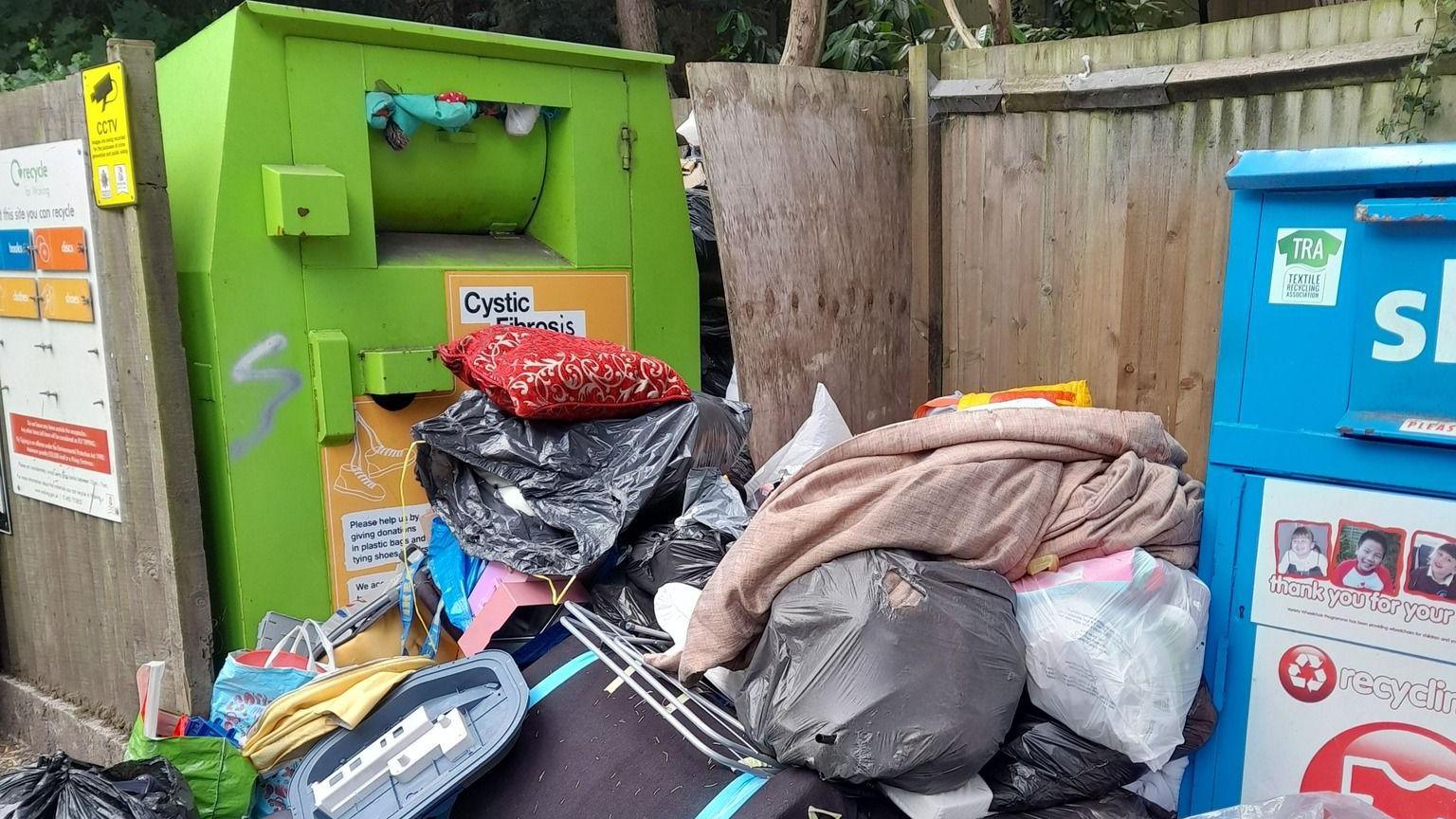 A pile of household waste, including black bags, full carrier bags and other items. The pile is between a large green recycling bin and a similar blue bin, and surrounded by wooden fencing.