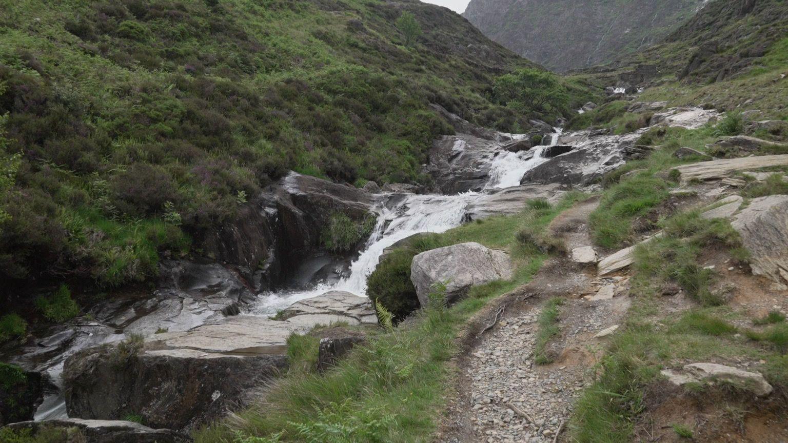A stream cascades down a series of rocks alongside a walking path. The bank slopes upwards on the left hand side.