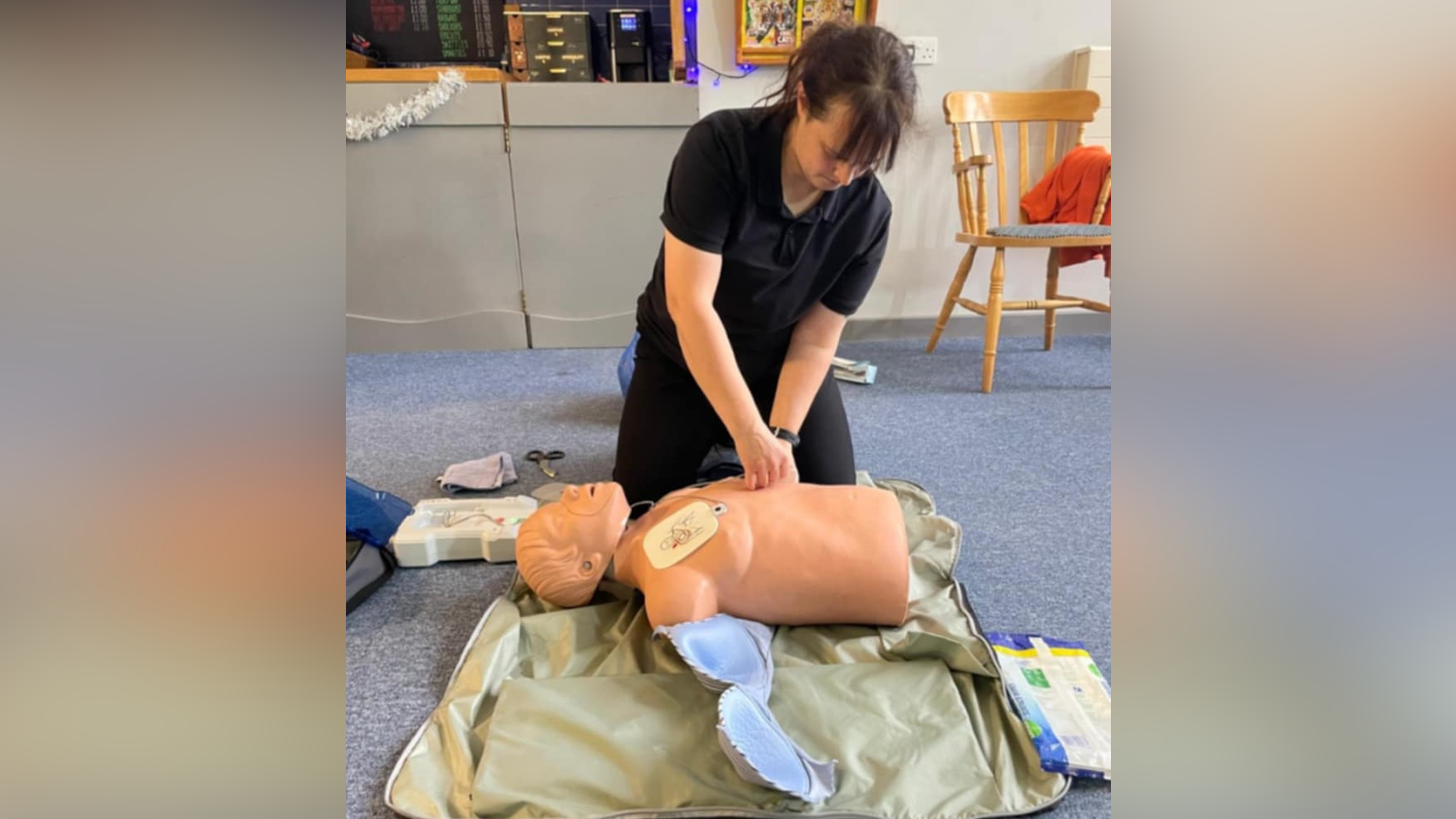 A woman is kneeling on the floor over a CPR dummy preparing to do a demonstration. She has dark hair with a fringe that is tied back. She is wearing a black T-shirt with black trousers and has her hands resting on the dummy's torso