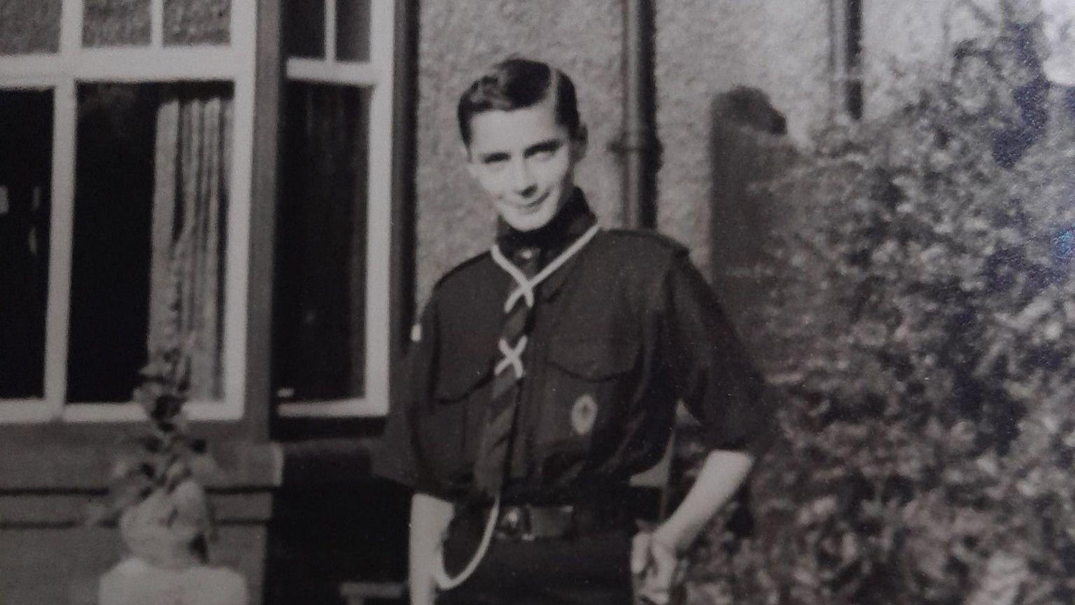 A black and white photograph of Geoffrey Crawford standing next to a shrub outside of a house. His short dark hair is combed to the side. He is wearing a dark shirt, trousers and a tie.