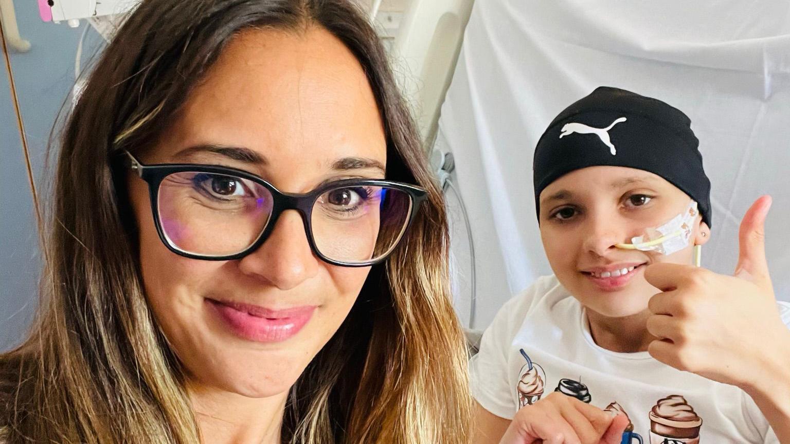 Lydia wearing glasses and smiling at the camera. She is taking a selfie in hospital with her daughter Amelie, who is laying in bed wearing a Slazenger cap with a tube up her nose. Amelie is smiling and giving a thumbs up to the camera. 