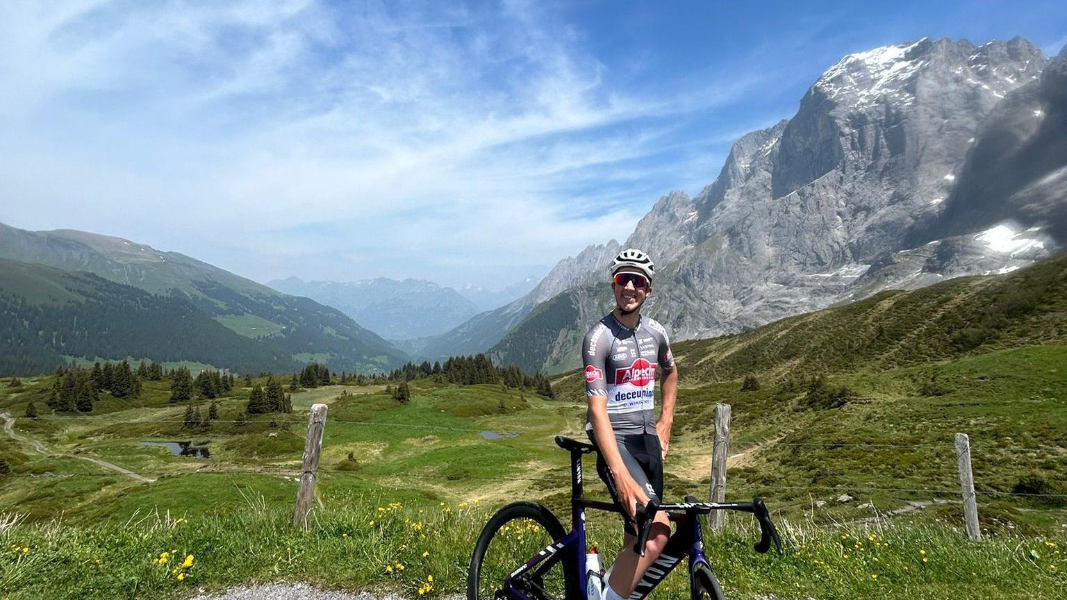 Jente Michels is smiling at the camera in a helmet and cycling gear. He is posing on his bike with mountain slopes towering behind him.