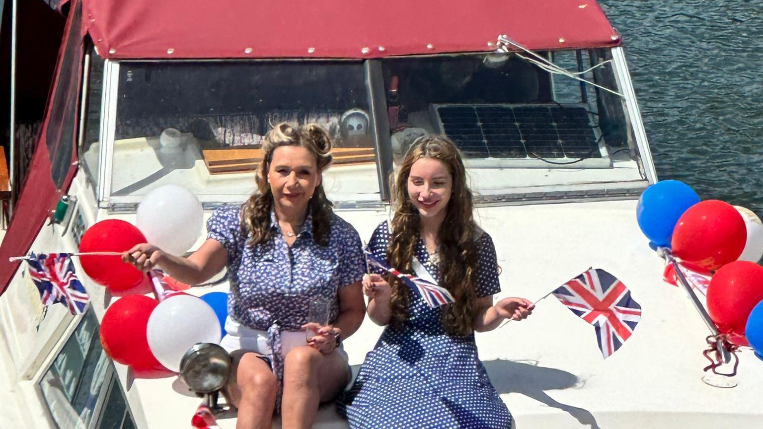 Two women in 40's style outfits and waving Union Jack flags to mark D-Day sit on a boat