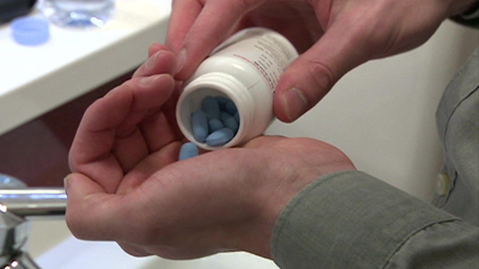 File image showing a man tipping a small white plastic bottle containing blue PrEP pills until one falls into his hand. A bathroom sink and blue bottle of water can be seen in the background 