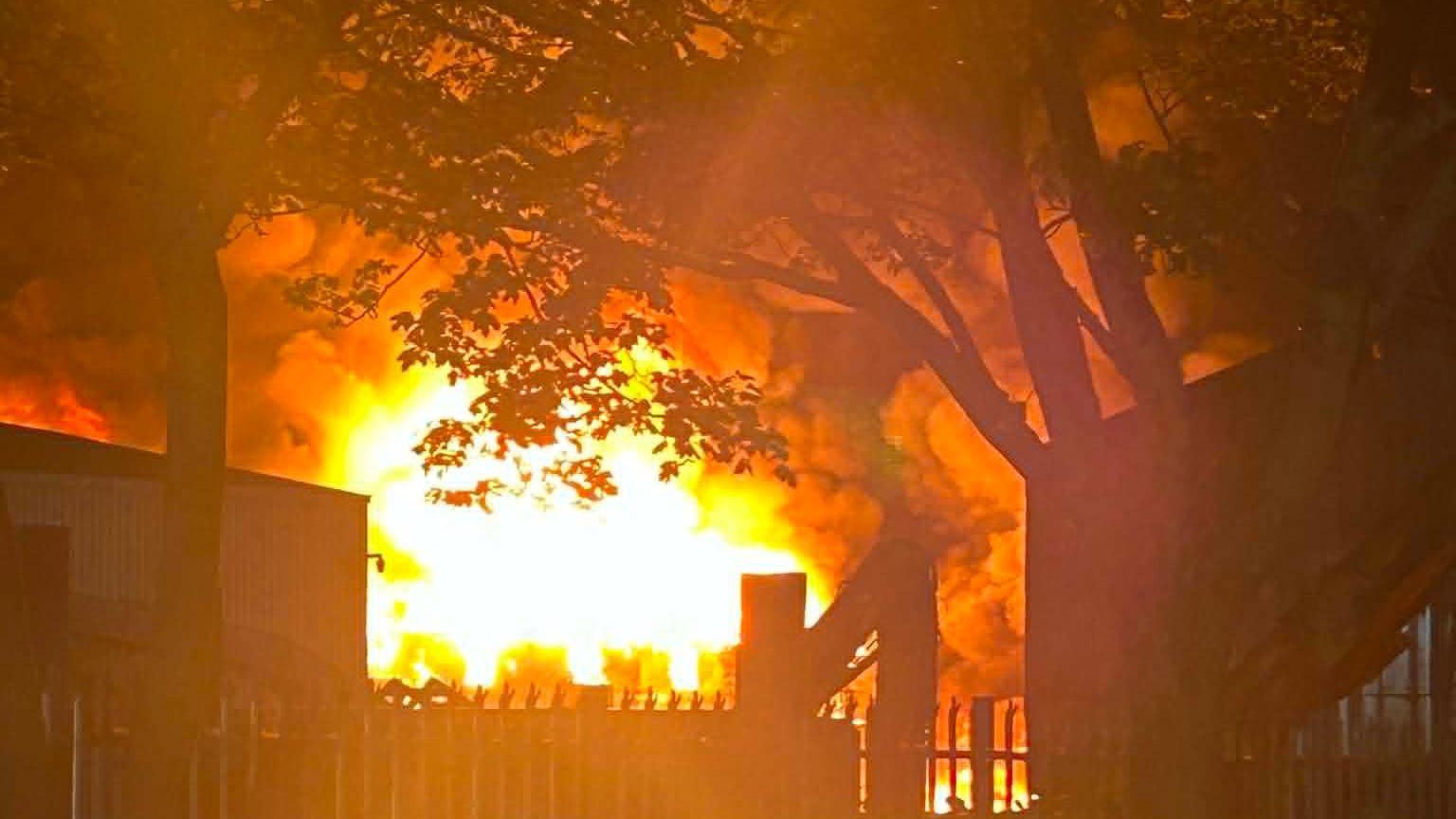 Flames coming from a building, showing a tree and fencing in front of it. The image is dark, but the flames are bright. There is also smoke in the air. 