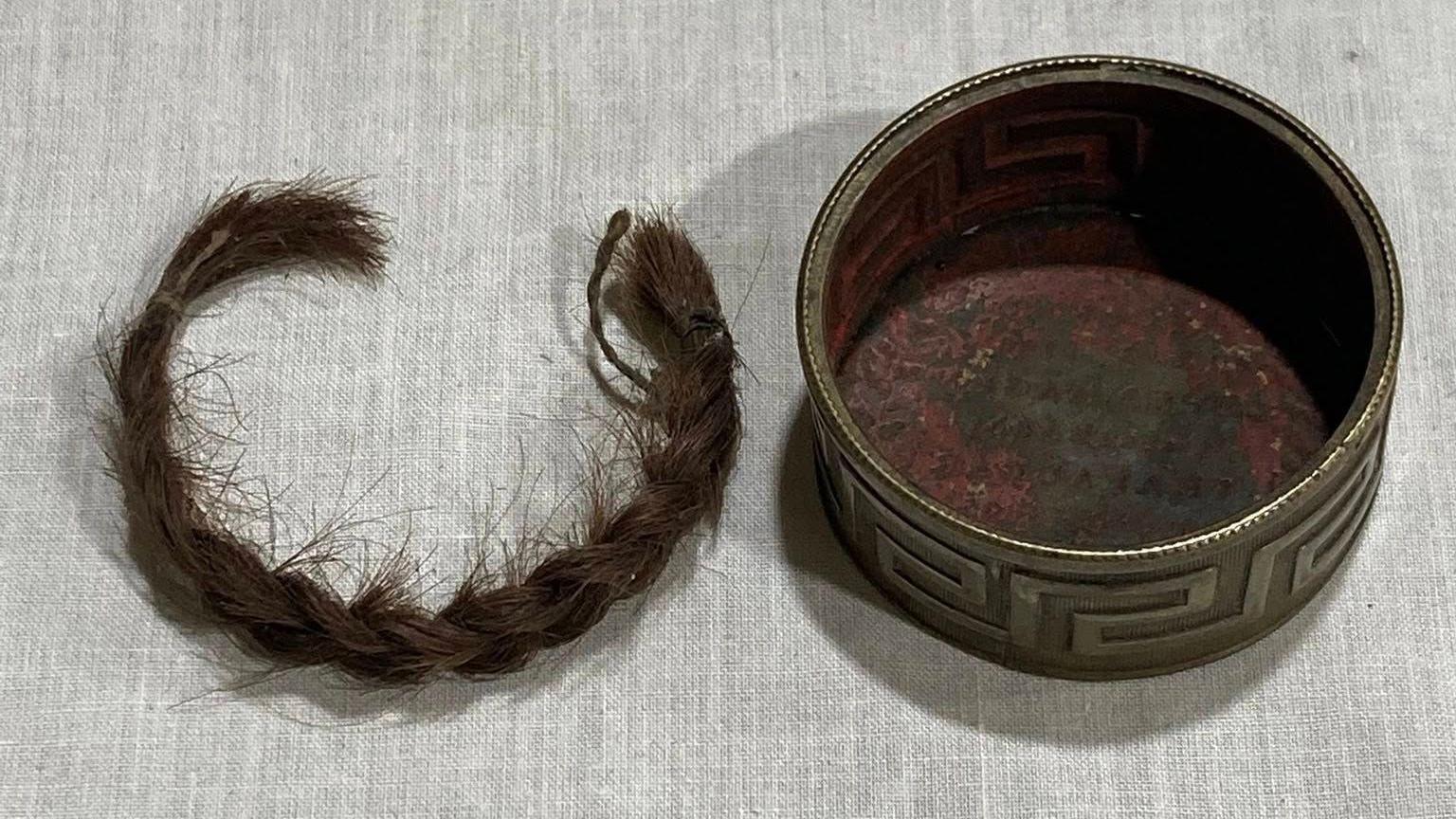 A dark brown plaited lock of hair is on a piece of cloth on the left hand side of a small gold box.