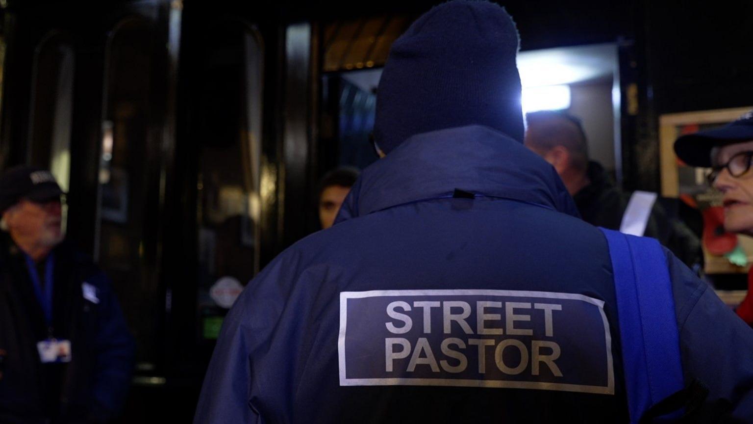 A person in a blue jacket with "street pastor" on the back, talking to a group of people at night in Stafford town centre.