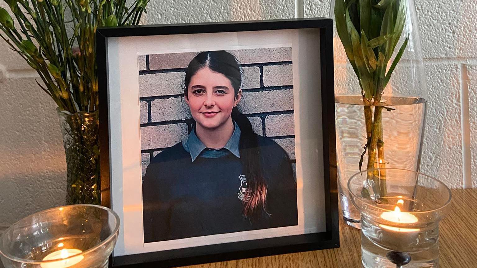 A framed photograph of Lauren who is wearing a navy school jumper and has a grey shirt collar. She has very long brown hair in a ponytail that is coming over her shoulder.