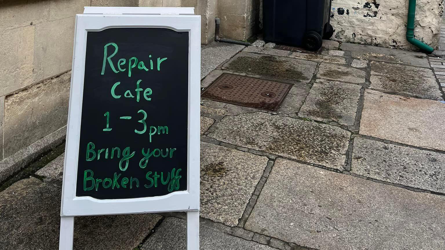 A chalk A-board sign outside a stone building that says 'Repair cafe 1-3pm, bring your broken stuff'