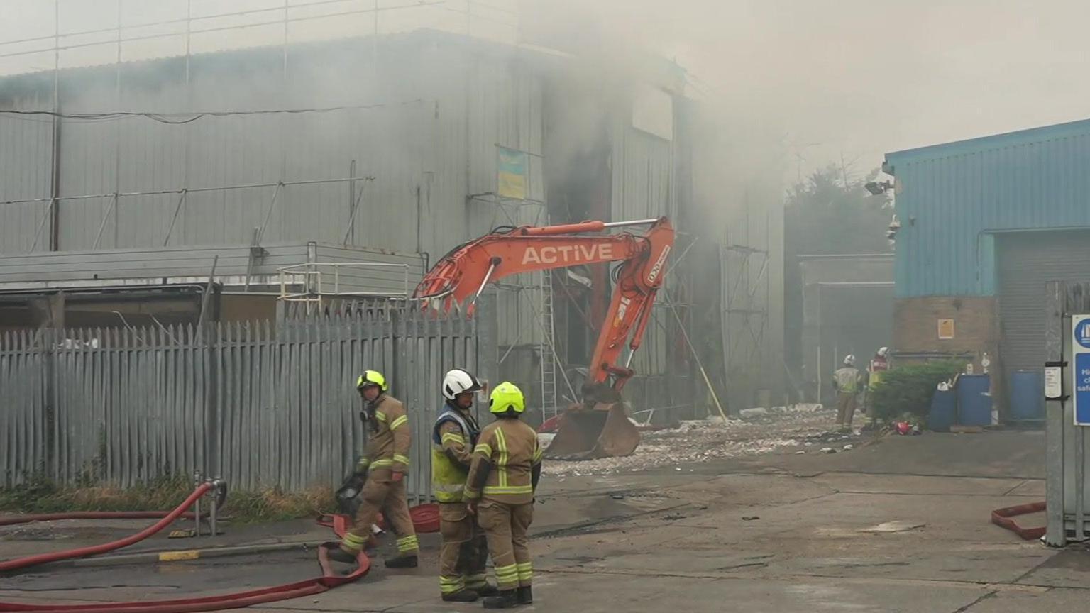 Firemen are standing outside of a silver warehouse. There is smoke coming out of the building and an orange excavator out the front. 