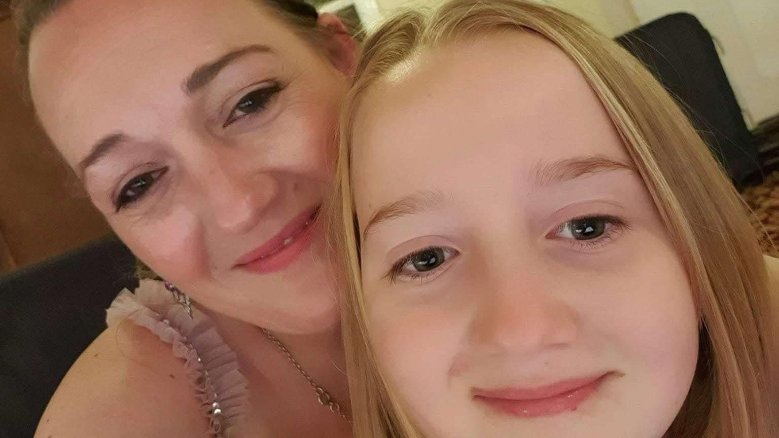 Kristine Reilly-Blake is smiling over her daughters shoulder. You can see the shoulder strap of her pink dress and her hair is tied back. Her daughter has straight blonde hair and is smiling at the camera 