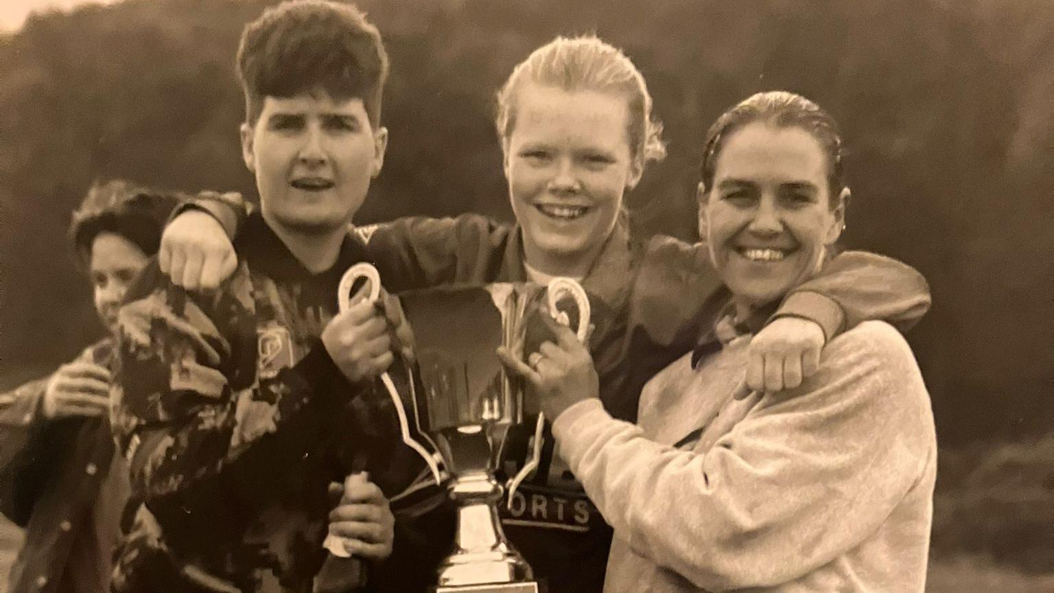 Marie Collins, Jo Cartwright and Elaine Vallack hold a trophy on a football pitch.