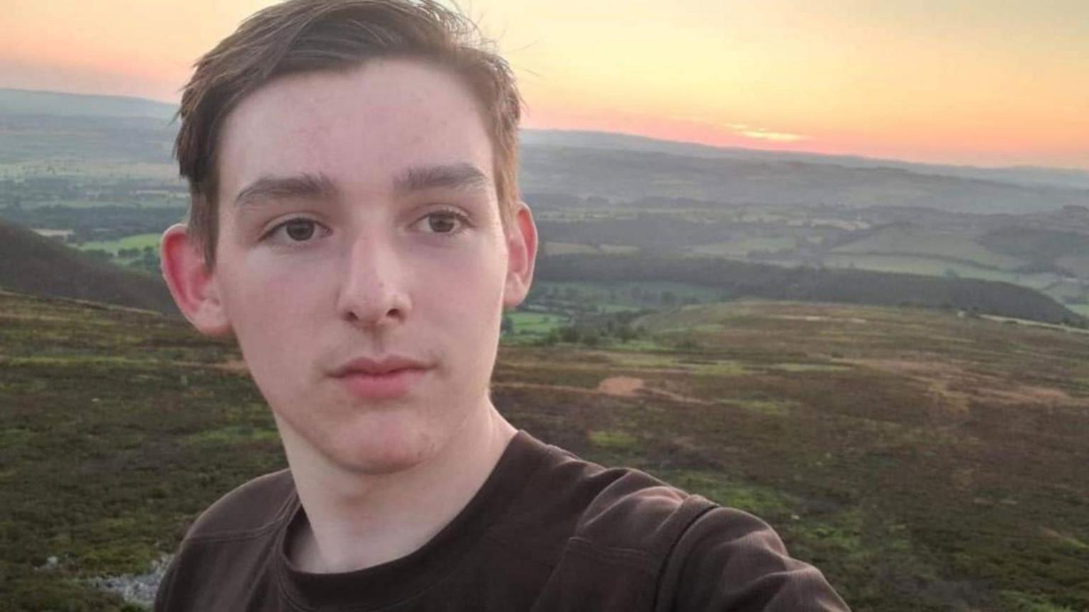 A young man with brown hair and a dark tshirt, standing on a hill with the sun behind him