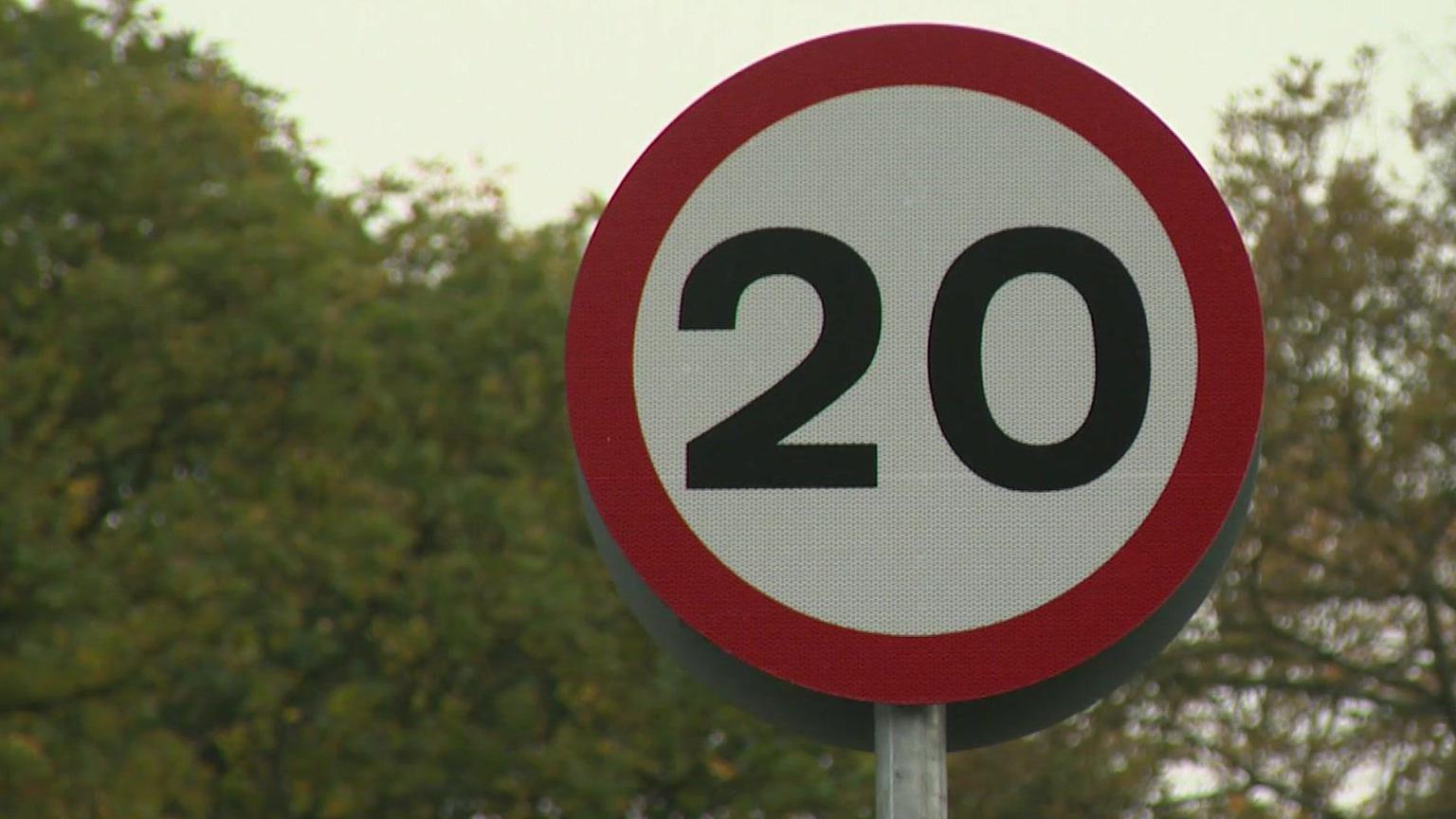 A close-up of a red circular road sign with 20 in black writing against a white background. There are trees behind the sign.