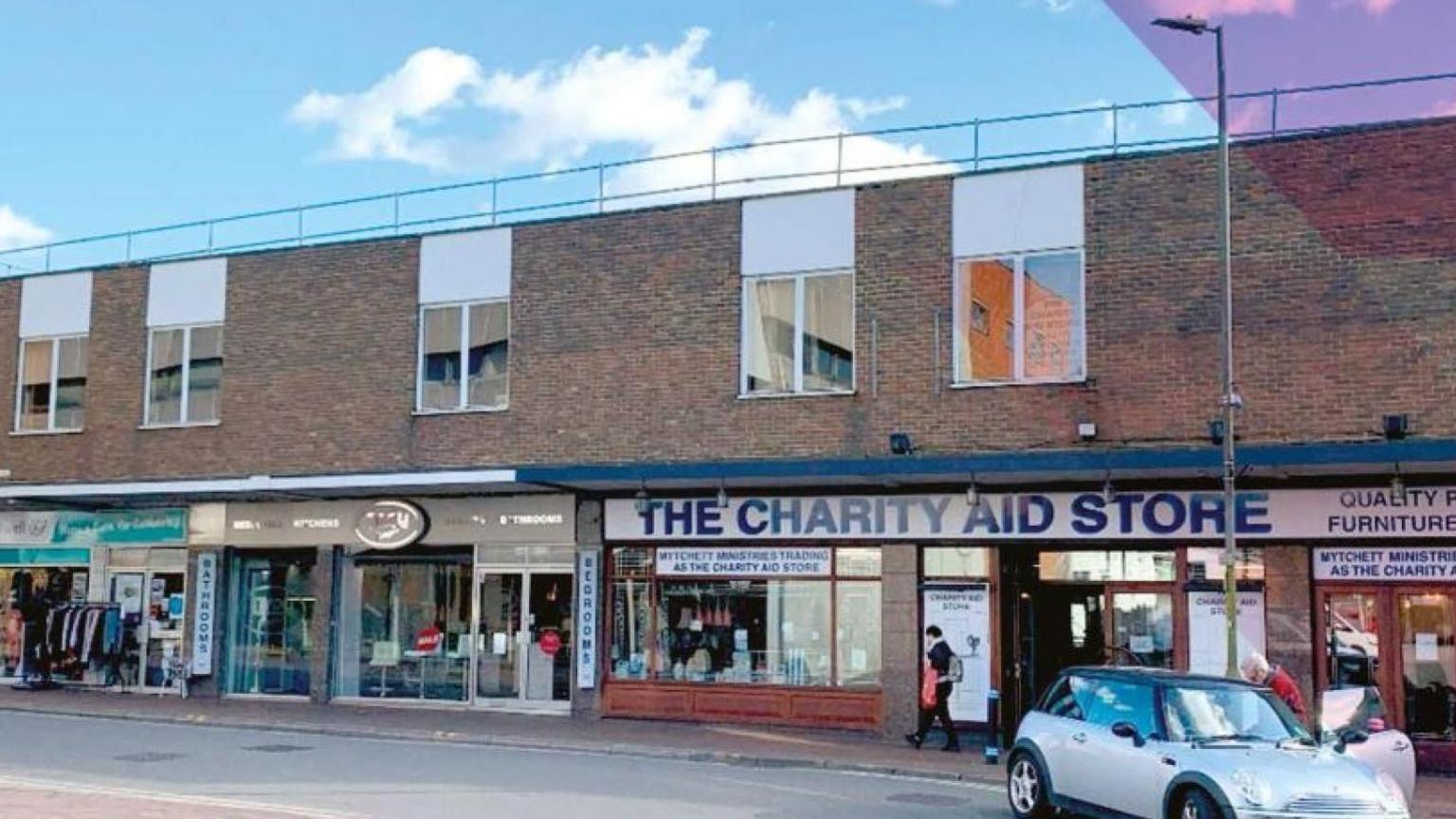 A row of shops in Camberley, including a charity shop. A mini is parked at the kerb and a pedestrian is walking past.