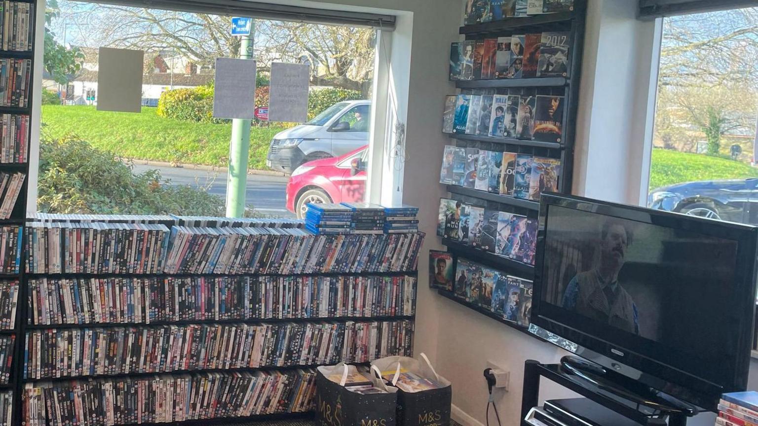 Shelves packed with DVDs on another wall of the shop. There is also a TV on another wall with a film playing.