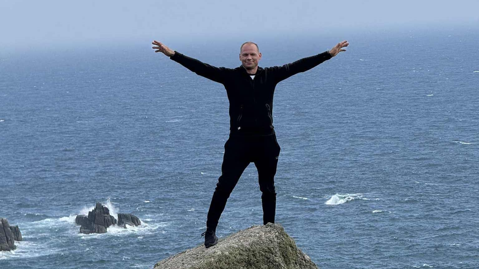 Tomas wearing a black jacket and black trousers- standing on a cliff with his arms spread out in the air - with the sea behind him