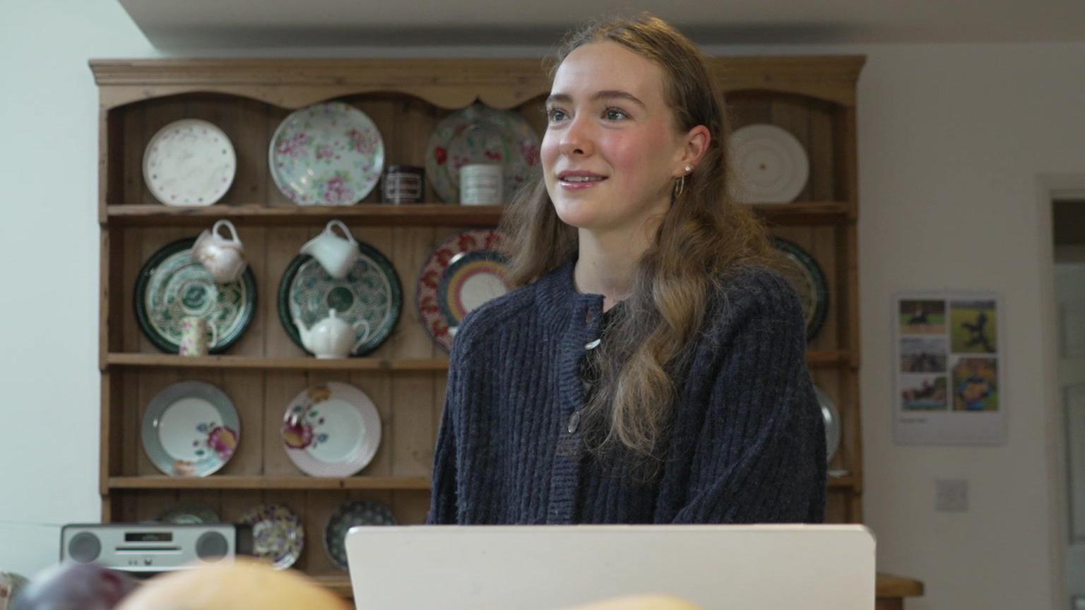 Lucy is stood at a dining table, her laptop in front of her. She wears a dark blue cardigan. Shelves of crockery are behind her.