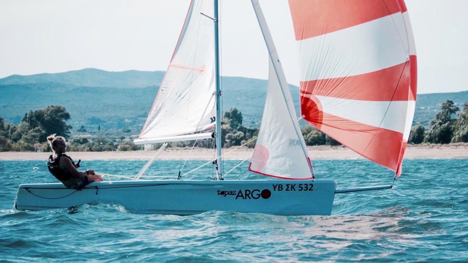 A young woman leans backwards off the back of a sailing dinghy. The Topaz Argo dinghy is white and she is sat down wearing a life jacket. The dignhy has two sails which are white and a spinnaker which is comprised of red and white horizontal stripes. The dinghy is on the water with a beach in the background. 